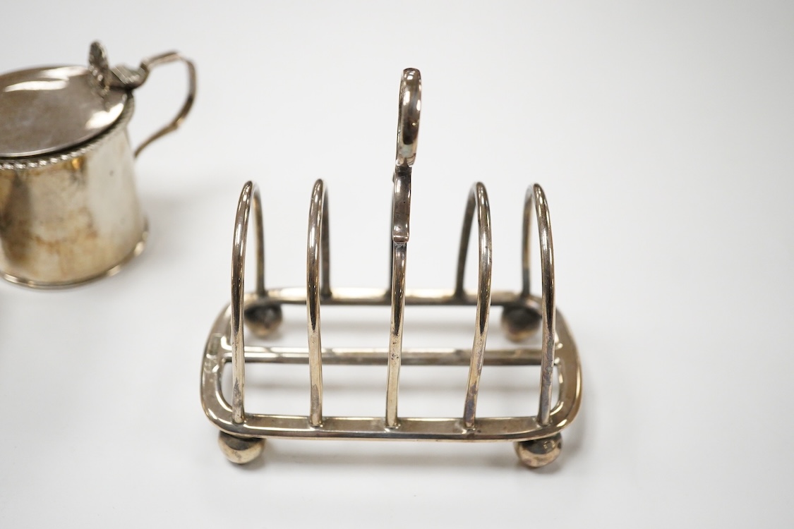 An Edwardian silver mug, on claw and hoof feet, William Hutton & Sons, London 1909, height 83mm, together with a silver butter shell, mustard pot and a silver five bar toastrack, 17oz.